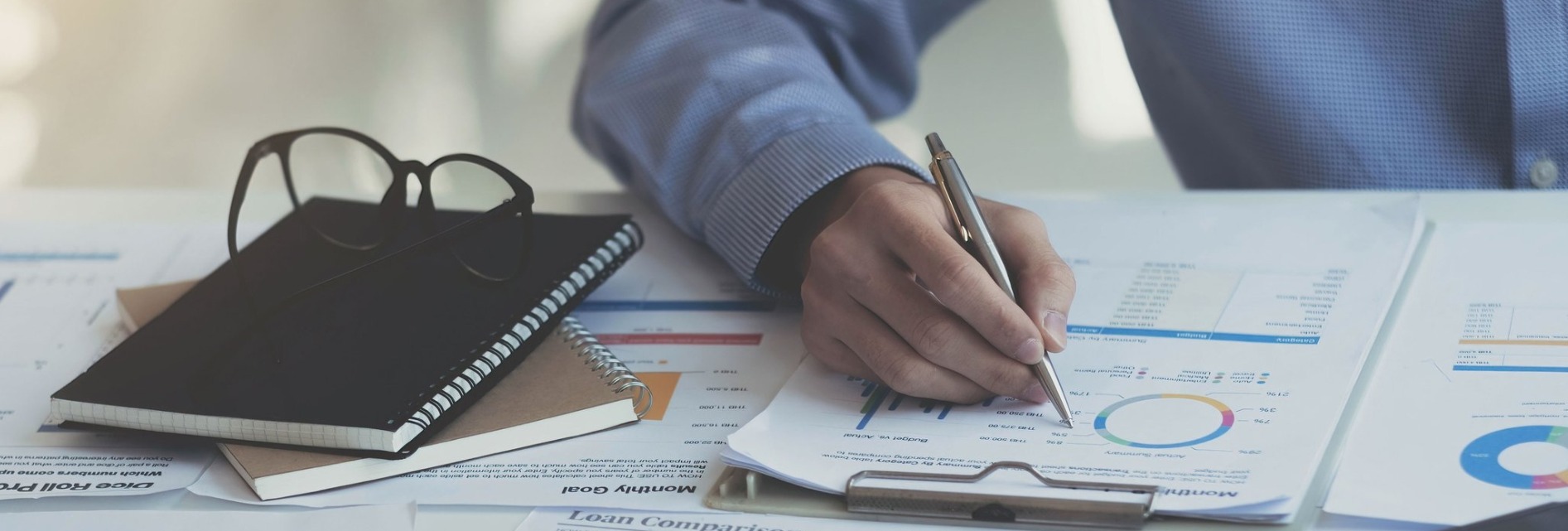 desktop with data charts, glasses and a hand holding pen 